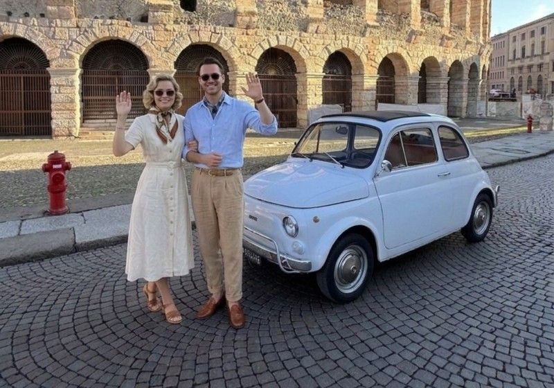 Una Fiat 500 bianca d'epoca ferma sul selciato davanti agli archi dell'Arena di Verona, con due passeggeri accanto
