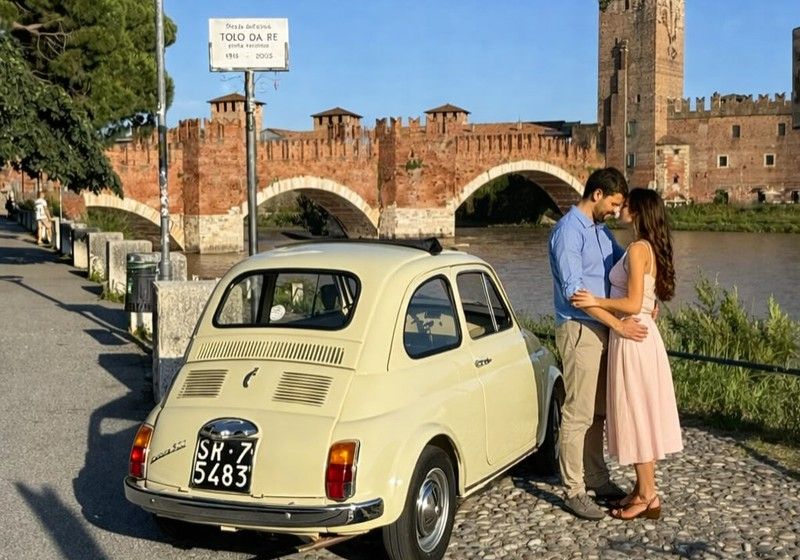 Una Fiat 500 avorio d'epoca vista da dietro davanti al ponte di Castelvecchio a Verona, con una coppia abbracciata accanto